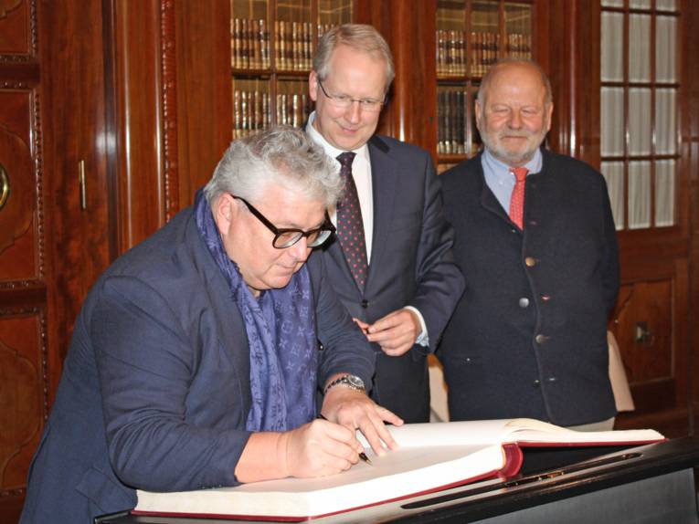 Drei Männer stehen hinter einem Stehpult mit einem großen Buch. einer der Männer schreibt in das Buch hinein. 