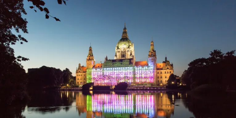 Bunt beleuchtetes Rathaus. Davor ein Teich. 