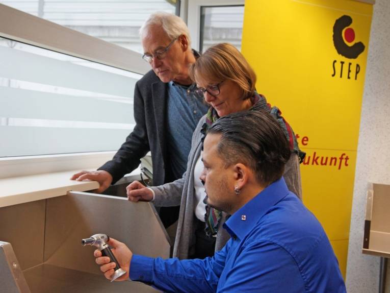 Ein Mann zeigt einer Frau und einem weiteren Mann ein technisches Gerät.