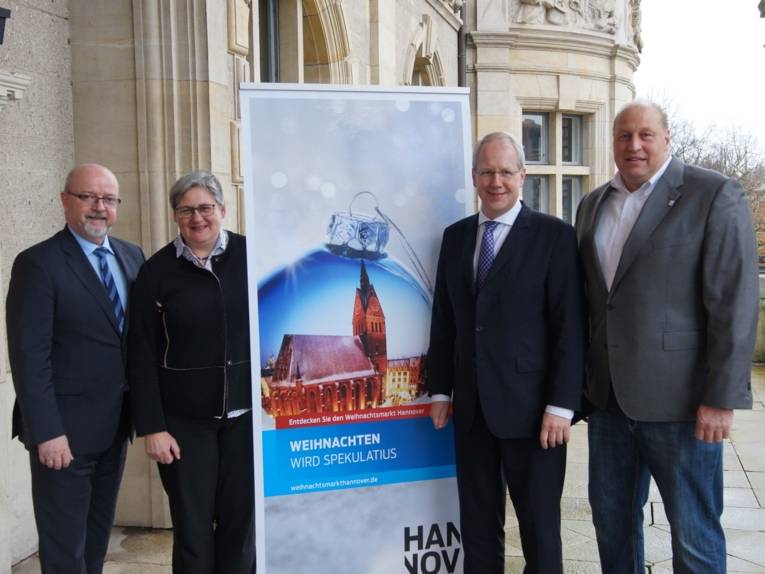 Drei Männer und eine Frau neben einem Werbebanner, auf dem eine Weihnachtskugel abgebildet ist. 