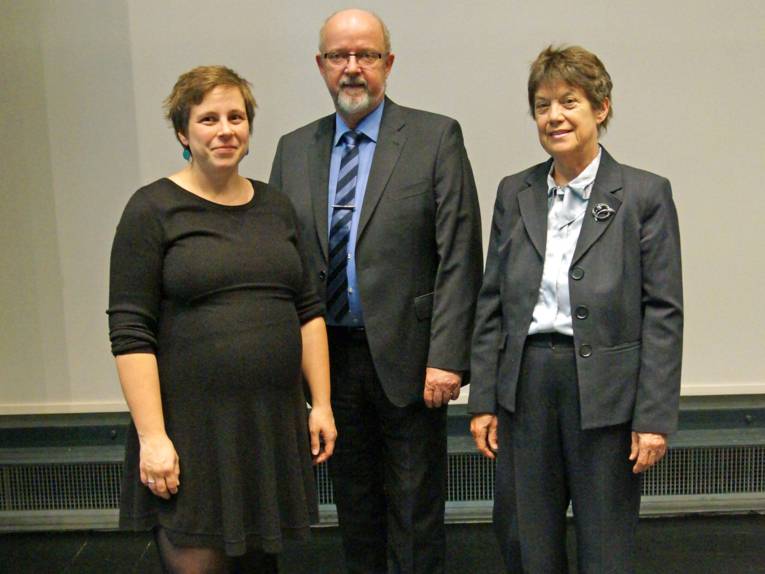 Zwei Frauen und ein Mann vor einer weißen Wand. 