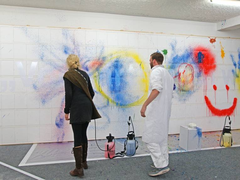 Eine Frau wirft einen Farbbeutel an eine weiße Wand, ein Mann im weißen Schutzanzug hantiert mit Farbsprühflaschen.