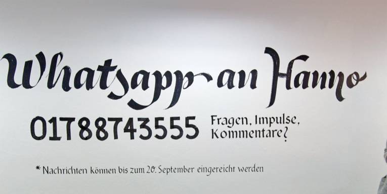 Auf eine weiße Wand ist folgende Nachricht gepinselt: Whatsapp an Hanno, 017887433555, Fragen, Impulse, Kommentare? Nachrichten können bis zum 20. September eingereicht werden