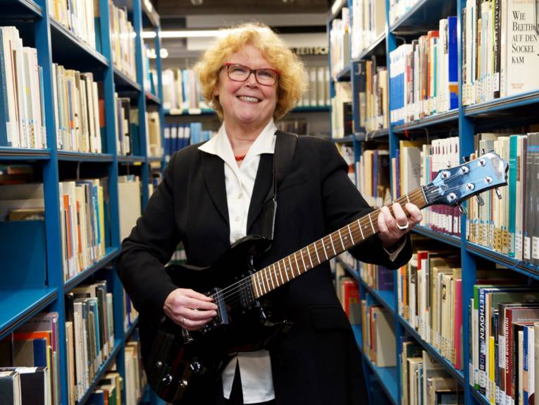 Frau mit einer Gitarre zwischen Bücherregalen. 