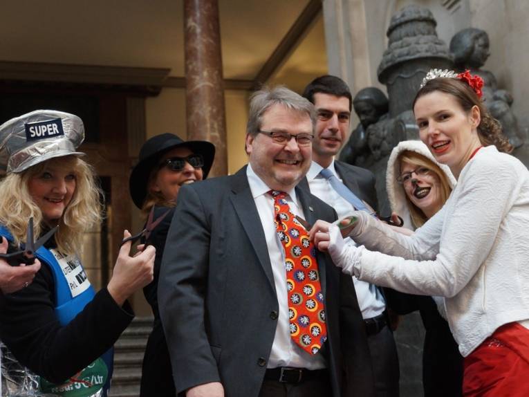 Zwei Männer mit Krawatten. Drumherum Frauen, die sie ihnen abschneiden. 