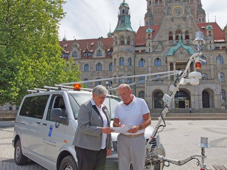 Eine Frau und ein Mann vor einem Messfahrzeug auf einem Platz. 