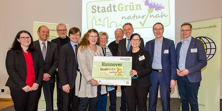 Gruppenfoto: Im Vordergrund halten Bürgermeisterin Regine Kramarek und Projektleiterin Annemarie Hische gemeinsam ein Schild mit dem Label "Hannover Stadtgrün naturnah", dahinter stehen weitere Personen, die sich freuen.