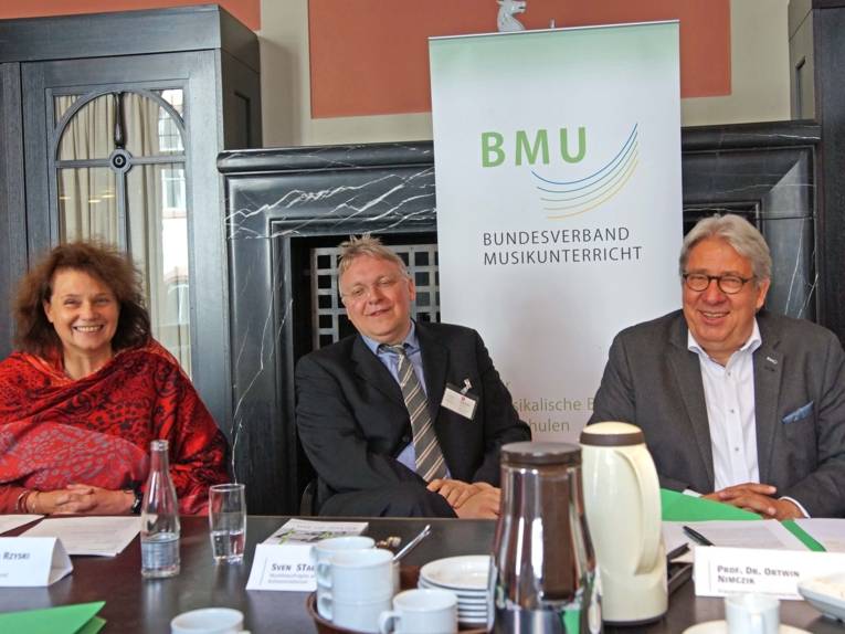 Eine Frau und zwei Männer hinter Tischen. 