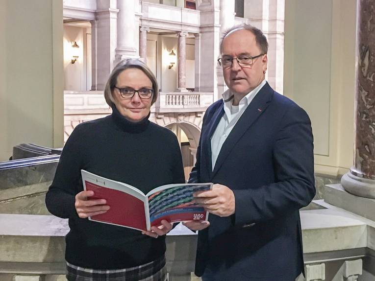 Eine Frau und ein Mann stehen an einer Balustrade, die Frau hält eine Broschüre in der Hand