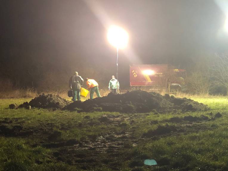 Drei Menschen um einen Erdhügel auf einem beleuchteten Feld. 