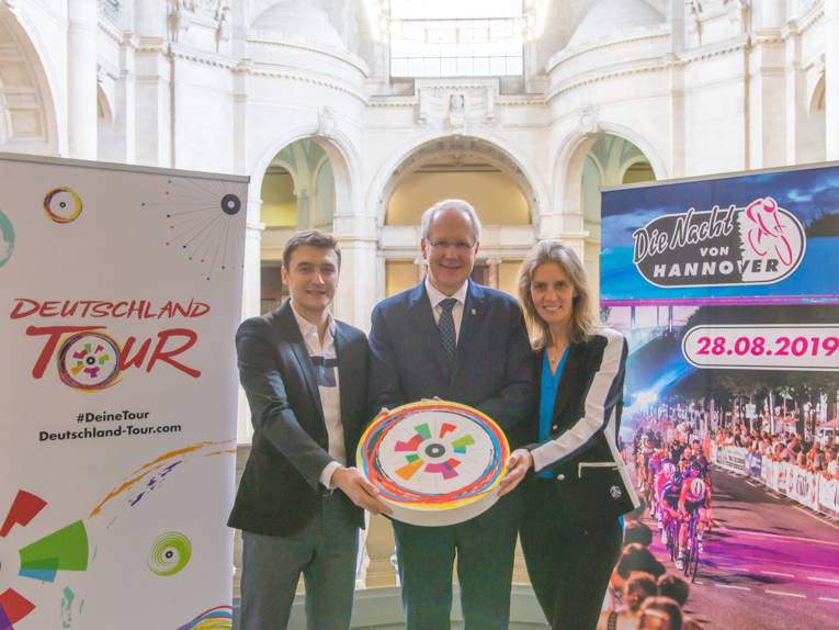 Drei Personen stehen neben zwei Roll-Ups, auf denen jeweils ein Radsport-Event angekündigt wird