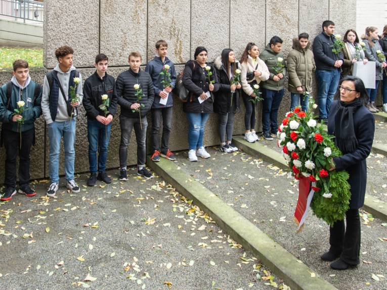 In einem Mahnmal stehen zahlreiche Personen, im Vordergrund eine Frau, die einen Kranz in den Händen hält