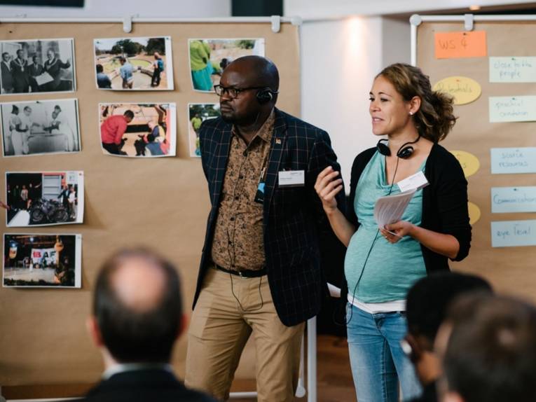 Ein Mann und eine Frau stehen vor zwei Stellwänden mit Fotos und Aushängen und präsentieren etwas.