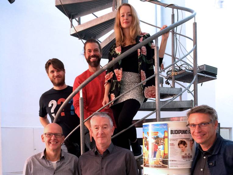 Sechs Personen posieren auf und vor einer Wendeltreppe, im Vordergrund rechts eine Säule mit Filmplakaten.
