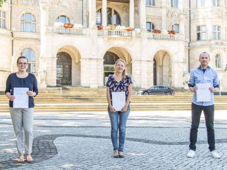 Drei junge Menschen vor einem Rathaus. Sie halten ein Abschlusszeugnis. 
