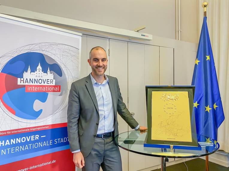 Belit Onay steht an einem Tisch, auf dem die Ehrenplakette des Europarates steht, daneben die Flagge der EU und ein Aufsteller mit der Aufschrift "Hannover – Internationale Stadt"