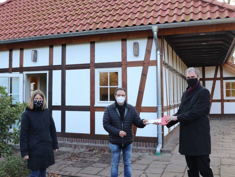 Drei Personen vor einem Fachwerkhaus in einem Park. Ein Herr übergibt einem anderen einen Schlüssel. 