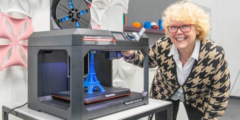 Eine Frau steht lächelnd neben einem 3D-Drucker, der gerade ein Modell des Eiffelturms produziert