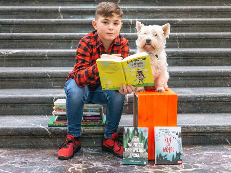 Ein Junge sitzt auf einem Stapel Bücher an einer Treppe, neben ihm sitzt auf einer Kiste ein Hund