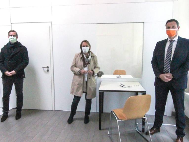 Drei Personen mit Mund-Nasen-Schutz in einem Raum. 