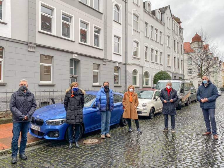 Sechs Personen stehen mit Masken und Abstand zueinander auf einer Straße vor einem Haus