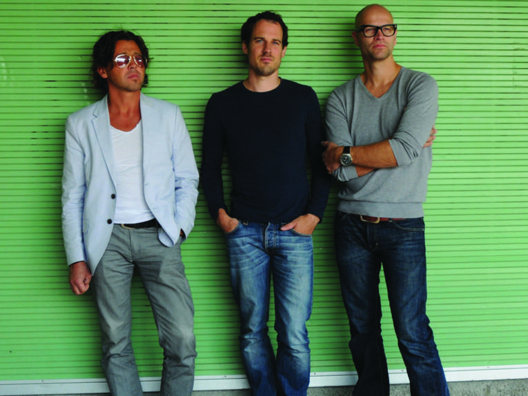 Drei Männer vor einer grünen Wand. 