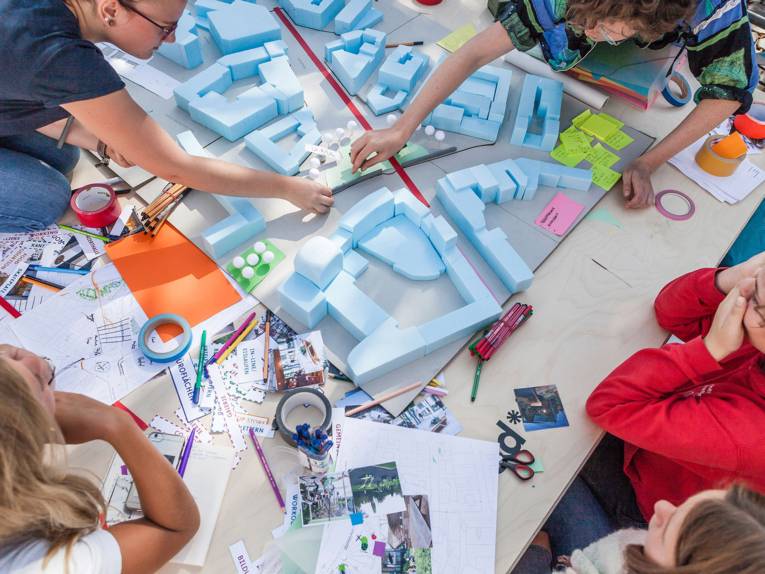 Fünf junge Leute haben sich um einen Tisch mit einem Papier-Modell des Steintorplatzes versammelt und schieben Elemente des Modells hin und her. Auf dem Tisch liegen zahlreiche Utensilien wie Stifte, Klebezettel, Scheren, Klebeband und ausgeschnittene Bilder und Papierstreifen.