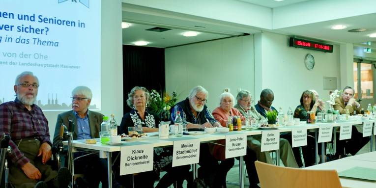 Die Mitglieder des Seniorenbeirats sitzen vorne in einer Reihe; dahinter ein Stück Projektionsfläche, auf der der Redner, Herr von der Ohe, zum Thema "Leben Seniorinnen und Senioren in Hannover sicher" angekündigt wird.