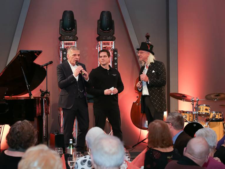 Drei Herren auf einer Bühne, daneben ein Klavier und ein Schlagzeug; vor der Bühne sitzt ein älteres Publikum.