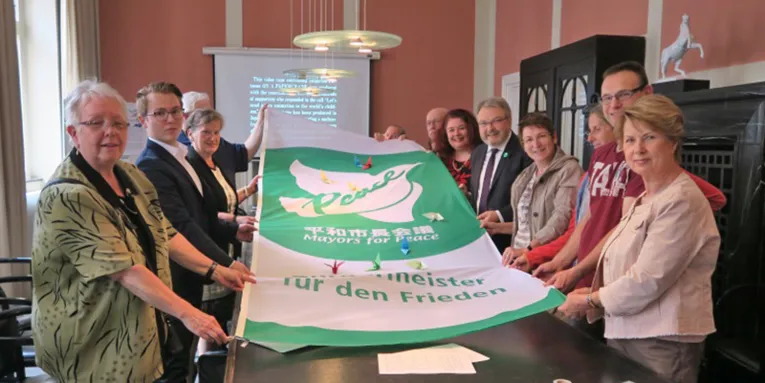 Über einen Tisch im Neuen Rathaus hält eine Gruppe von Menschen gemeinsam mit Bürgermeister Thomas Hermann die Mayors for Peace Fahne, die eine weiße Friedenstaube zeigt. Darauf befinden sich einige Papierkraniche.