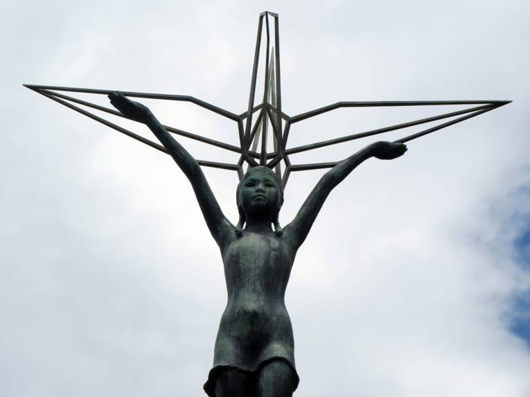 Die Statue eines Kindes mit ausgebreiteten Armen vor bewölktem Himmel.