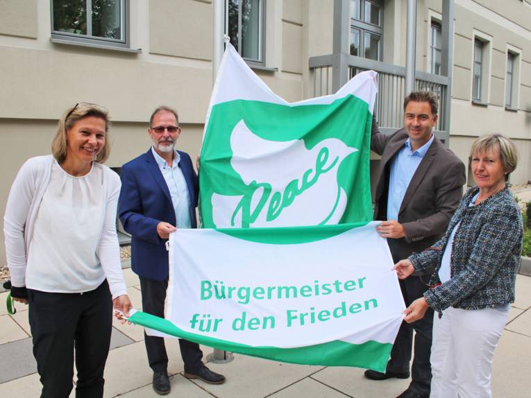 Vier Personen halten gemeinsam die "Bürgermeister für den Frieden" Flagge, ehe sie vor dem Rathaus von Königs Wusterhausen gehisst wird.