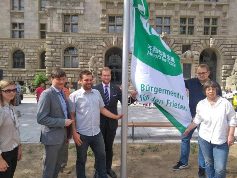 Sieben Personen stehen beim Hissen der Bürgermeister-für-den-Frieden-Fahne um einen Fahnenmast, der sich vor dem Neuen Rathaus Leipzig befindet.