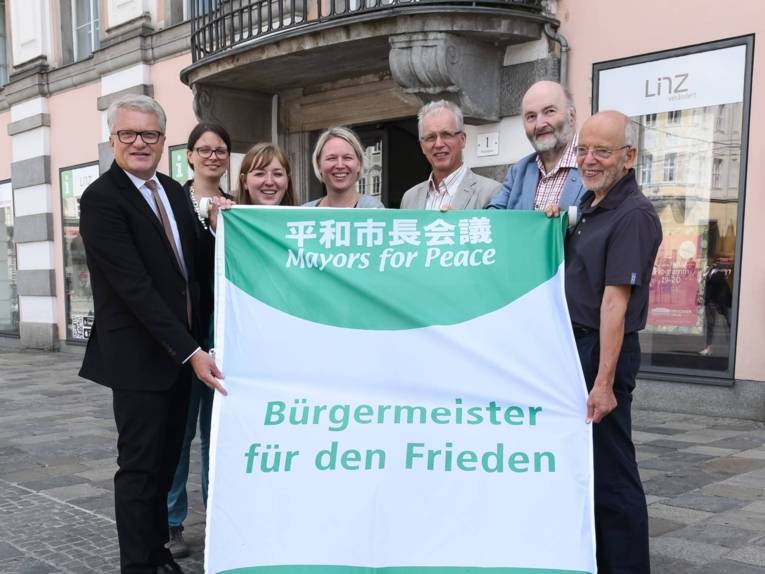 Bürgermeister Klaus Luger und sechs weitere Personen stehen vor dem Linzer Rathaus und zeigen die Flagge "Bürgermeister für den Frieden", bevor sie gehisst wird.