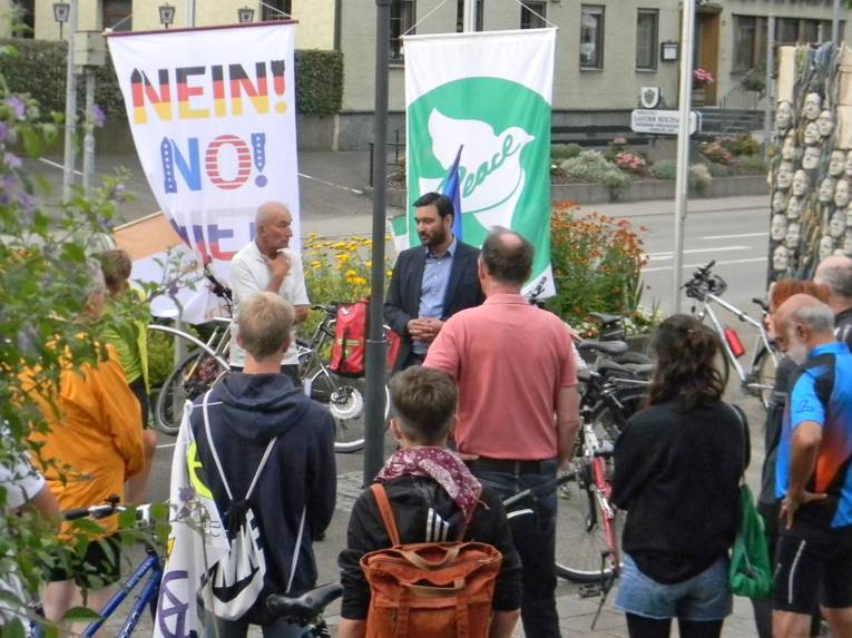 Mögglingens Bürgermeister und mehrere andere Personen stehen auf einem Platz in einem Kreis um zwei Flaggen: Die eine zeigt "Nein, No, Njet" als Zeichen gegen Atomwaffen und die andere die Friedenstaube mit der Aufschrift "Bürgermeister für den Frieden"