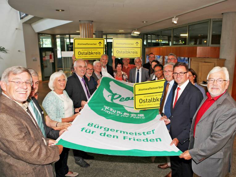 Zahlreiche Personen präsentieren die ausgebreitete Bürgermeister-für-den-Frieden-Flagge und gelbe Ortschilder des Ostalbkreises mit der Aufschrift "Atomwaffenverbot".