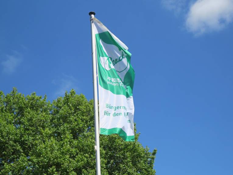 Die grün-weiße "Bürgermeister für den Frieden"-Flagge mit einer weißen Taube und dem Wort "Peace" weht im Wind vor blauem Himmel.