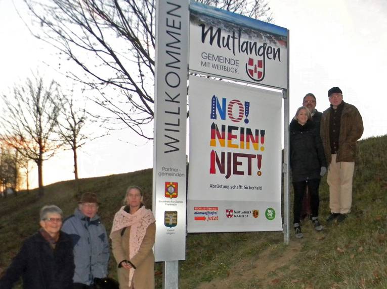 Drei Frauen und drei Männer stehen an einem Willkommensschild der Gemeinde Mutlangen, unter dem ein Banner mit der Aufschrift "No, Nein, Njet! Abrüstung schafft Sicherheit." angebracht wurde.