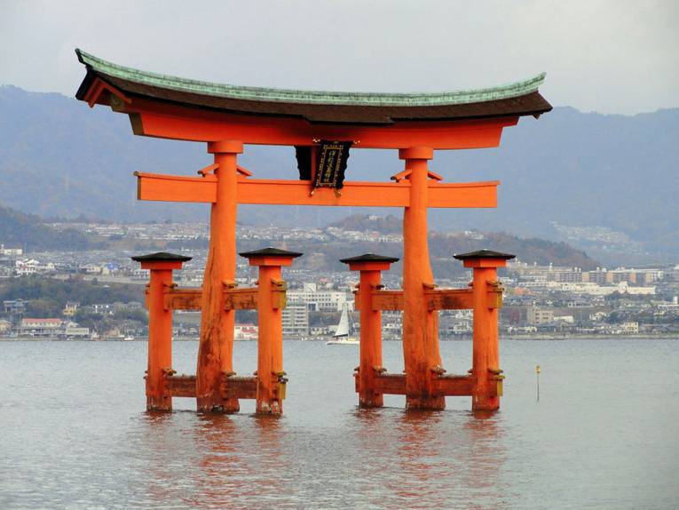 Das 16 m hohe Holztor in orange-roter Farbe mit geschwungenem Dach steht in einer Bucht Japans im Wasser und ist Teil des Itsukushima-Schreins.
