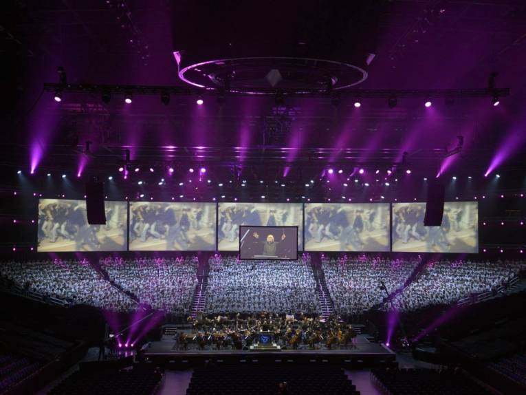 Ein Chor mit mehreren hundert Sänger*innen in einer großen Halle in violettem Licht.