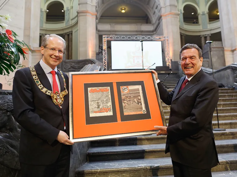 Stefan Schostok und Gerhard Schröder mit zwei eingerahmten historischen Fußballzeitschriften