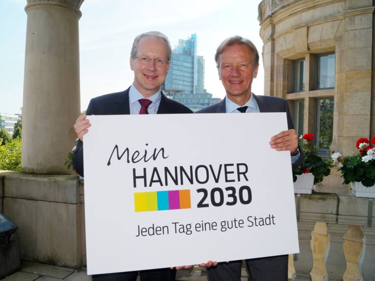 Stefan Schostok und Uwe Bodemann auf dem Rathausbalkon mit der Bild-Wort-Marke "Mein Hannover 2030 – Jeden Tag eine gute Stadt"