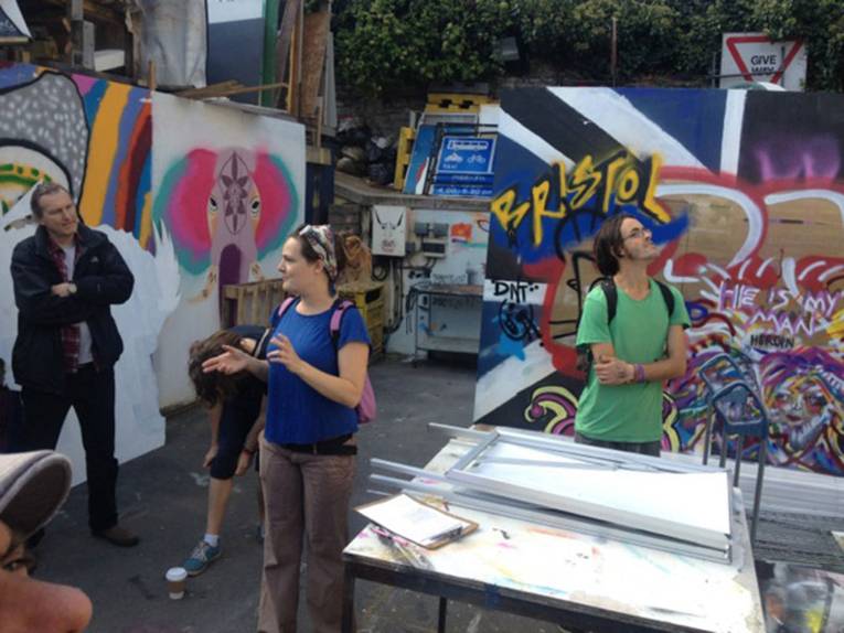 Eine Führerin erläutert den Zuhörenden das Gelände des Streetart Festivals. Im Hintergrund mit Graffiti besprühte Wände und Holzplatten.