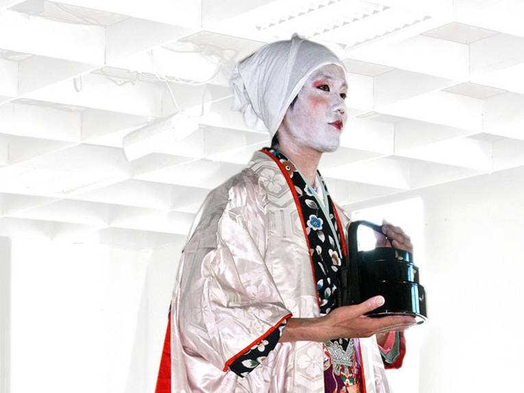 Eine traditionell nach japanischer Art geschminkte Person mit einer Teekanne in den Händen in einem weißen Raum