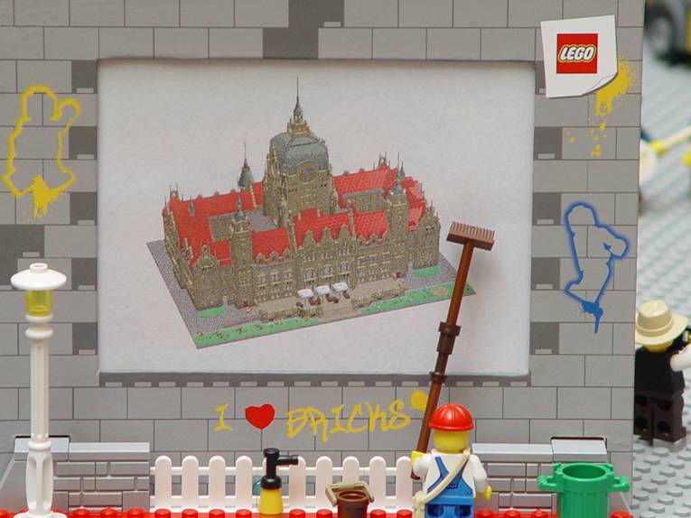 Eine Lego-Spielfigur steht vor einer Wand, auf der ein Modell des Neuen Rathauses abgebildet ist, wie es aus jetzt aus vielen Bausteinen entstehen soll