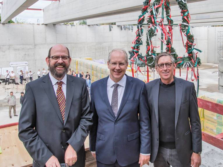 Drei Männer in einer Halle vor einem Kranz. 