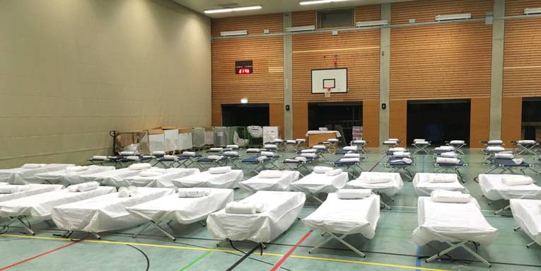 Mehrere Reihen von Feldbetten sind in einer Sporthalle aufgebaut