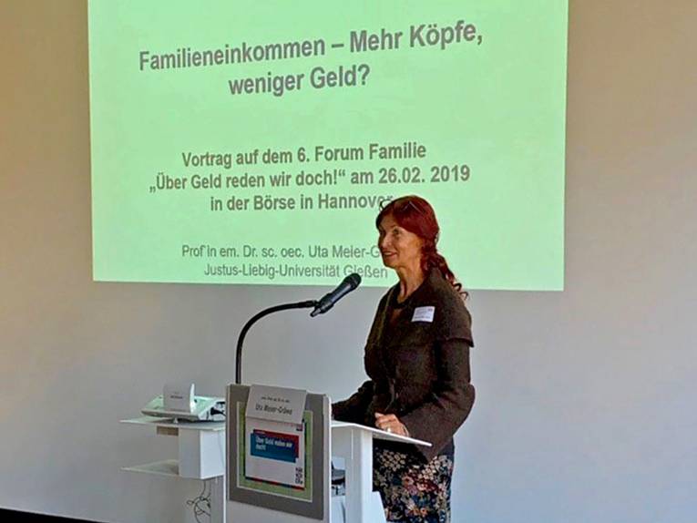 Prof. em. Dr. oec. Uta Meier-Gräwe am Redepult; dahinter ist der Beginn der Power Point Präsentation "Familieneinkommen: Mehr Köpfe, weniger Geld" an eine Leinwand projiziert.