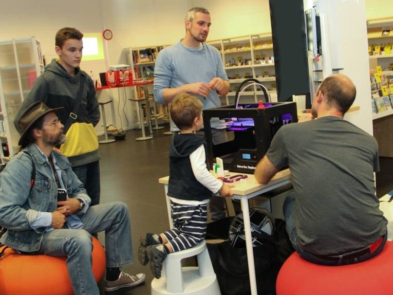 Ein 3D-Drucker in der Oststadtbibliothek, darum herum stehen, sitzen und knien fünf Jungen und Männer ganz unteschiedlichen Alters