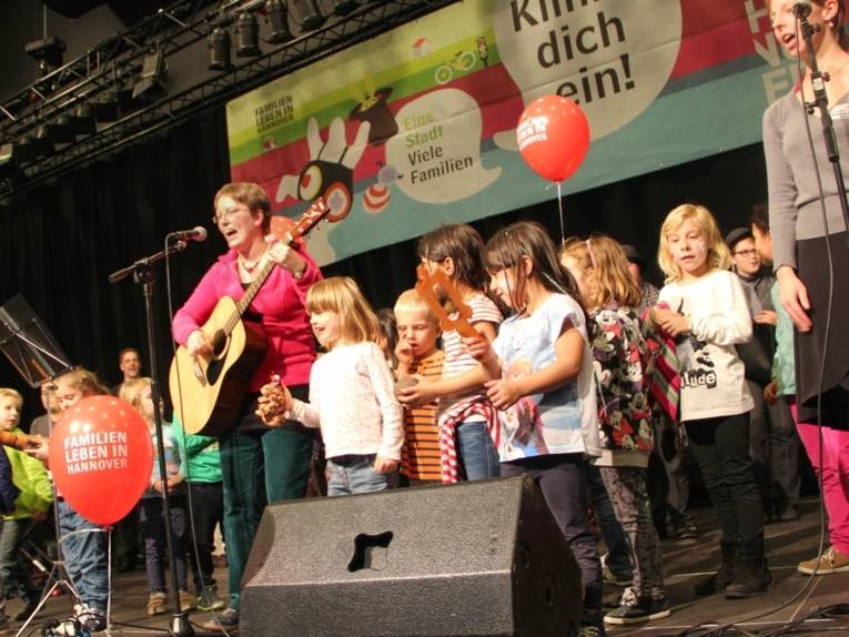 Zwei Frauen stehen auf einer Bühne und singen, eine von ihnen spielt Gitarre. Zwischen und neben ihnen stehen viele Kinder, von denen einige ein Rhytmusinstrument in der Hand haben. Im Hintergrund sieht man weitere Akteure des Familiensonntages.